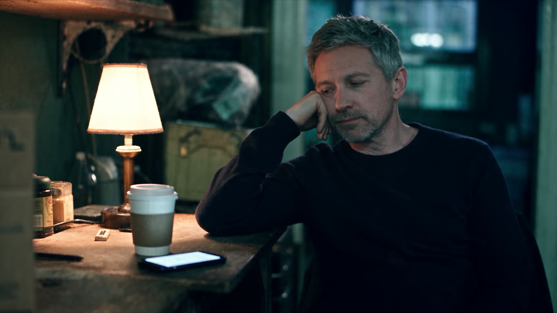 A man rests his head on his hand at a lamp-lit desk at night, his phone beside a coffee cup.