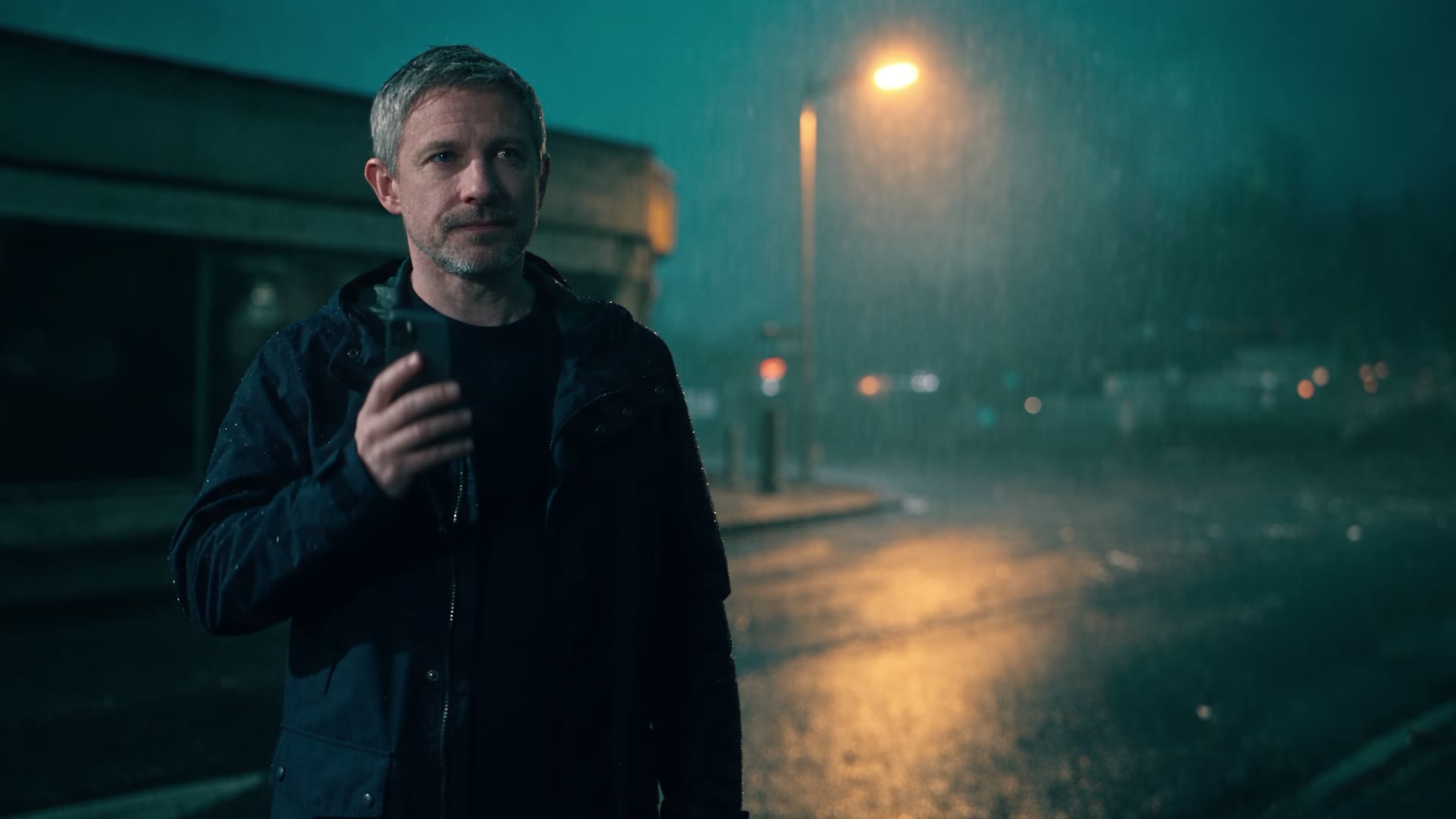 A man in a dark jacket stands on a wet street at night under a streetlight, his phone in his hand.